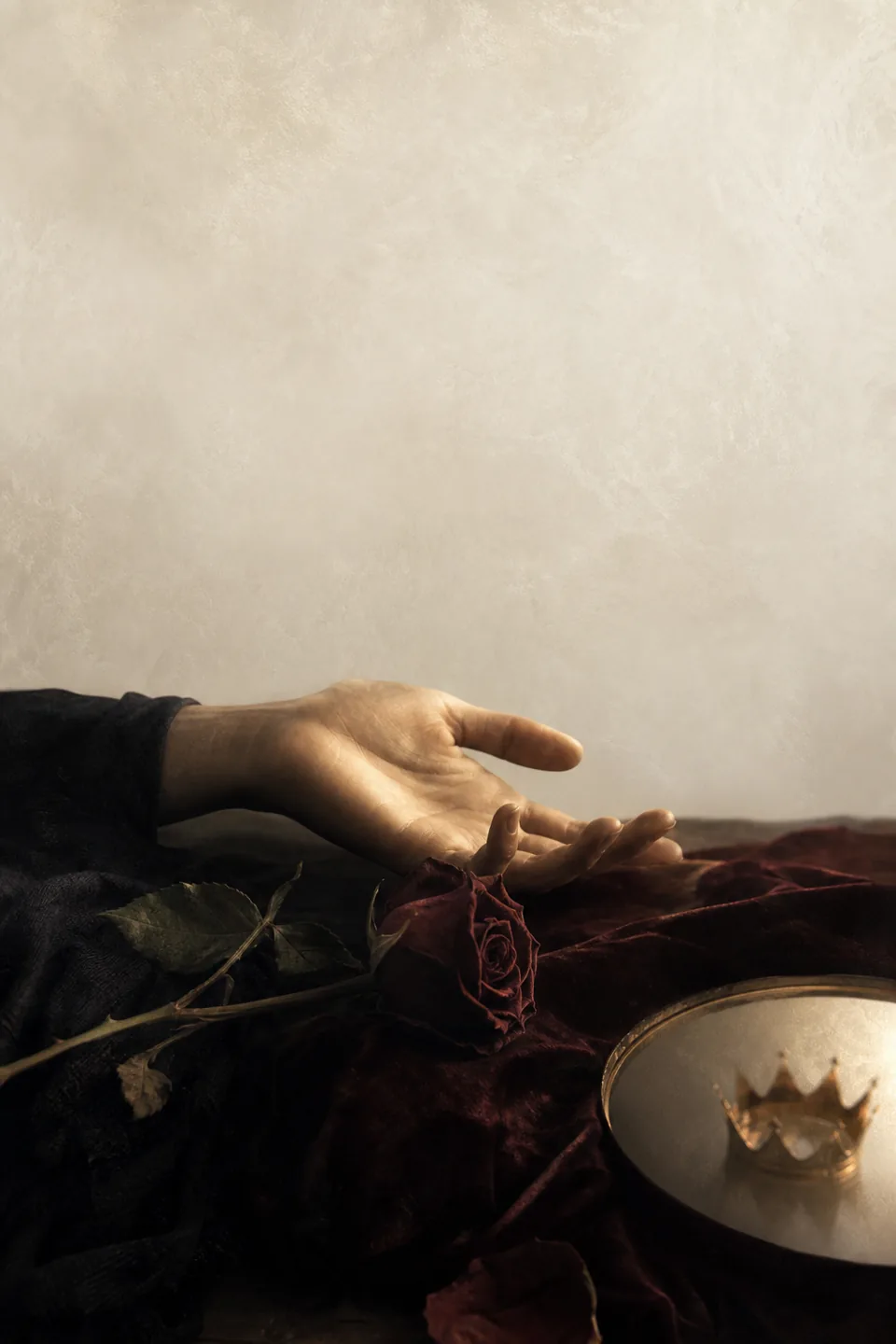 An open hand beside a dark red rose, with a faint crown-shaped reflection in soft window light.