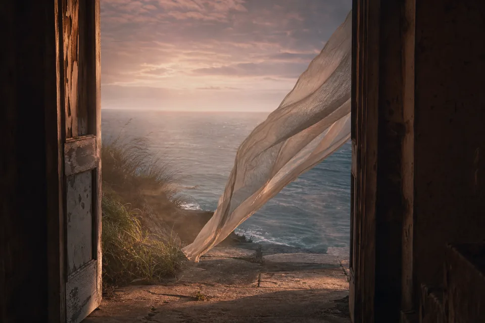 A loose veil carried by wind through an open stone doorway toward a quiet sea at dawn.